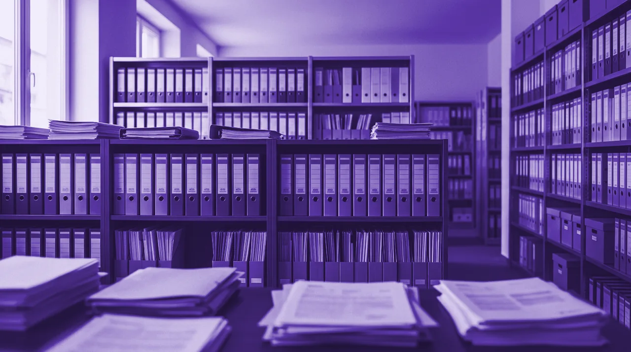 Tradycyjne archiwum dokumentów: regały pełne segregatorów i teczek oraz stosy papierowych akt.